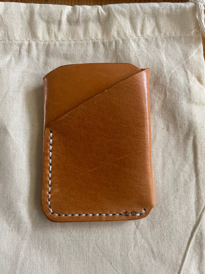 Handcrafted tan leather card holder with dual pockets, minimalist design, and hand-stitched edges on a neutral fabric background.